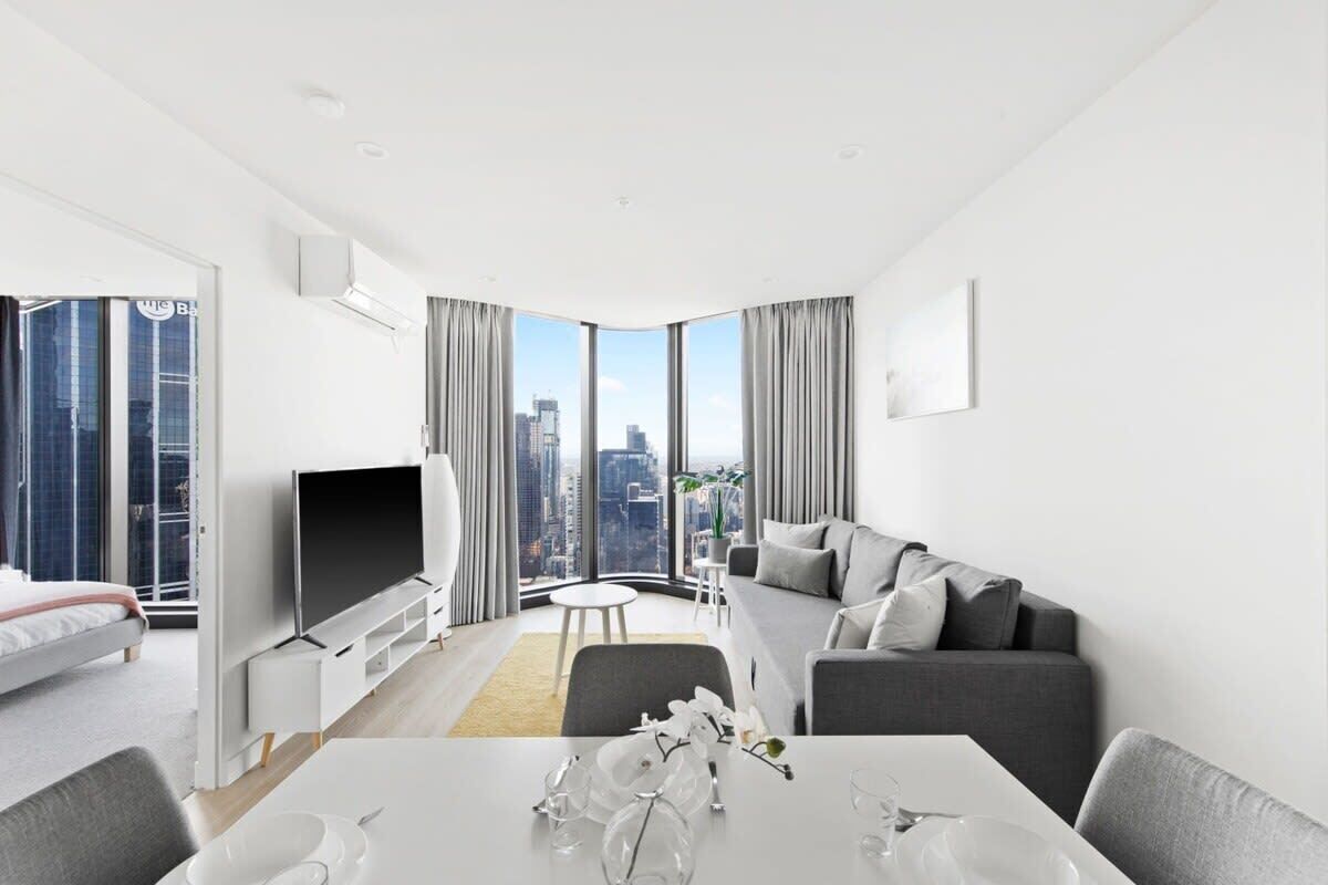 Ein modernes Hotelzimmer mit großem Fenster und Blick auf die Stadt, einem Flachbildfernseher, einem grauen Sofa, einem Esstisch mit weißer Dekoration sowie einem Bett mit weißem Kopfteil.