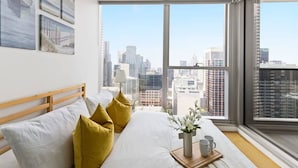 Een moderne slaapkamer met een groot raam, een bed met witte beddengoed en gele kussens, en uitzicht op een stadsbeeld.