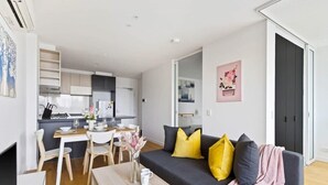 Een modern appartement met een eethoek, een bank met geel en grijze kussens, en een keuken op de achtergrond.