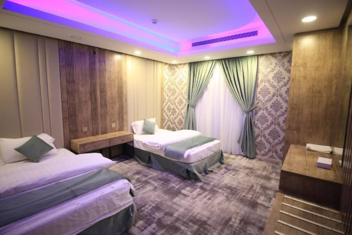 Habitación de hotel con dos camas, cabecero de madera, mesita de noche, tocador con espejo y papel tapiz estampado.
