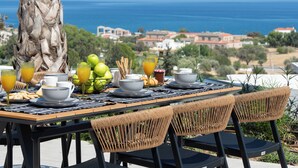 A table set for breakfast with a view of the sea, including plates, glasses, and a bowl of fruit.