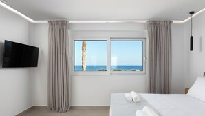 A modern hotel room with a large bed, two windows with curtains, a flat-screen TV, and a view of the sea.