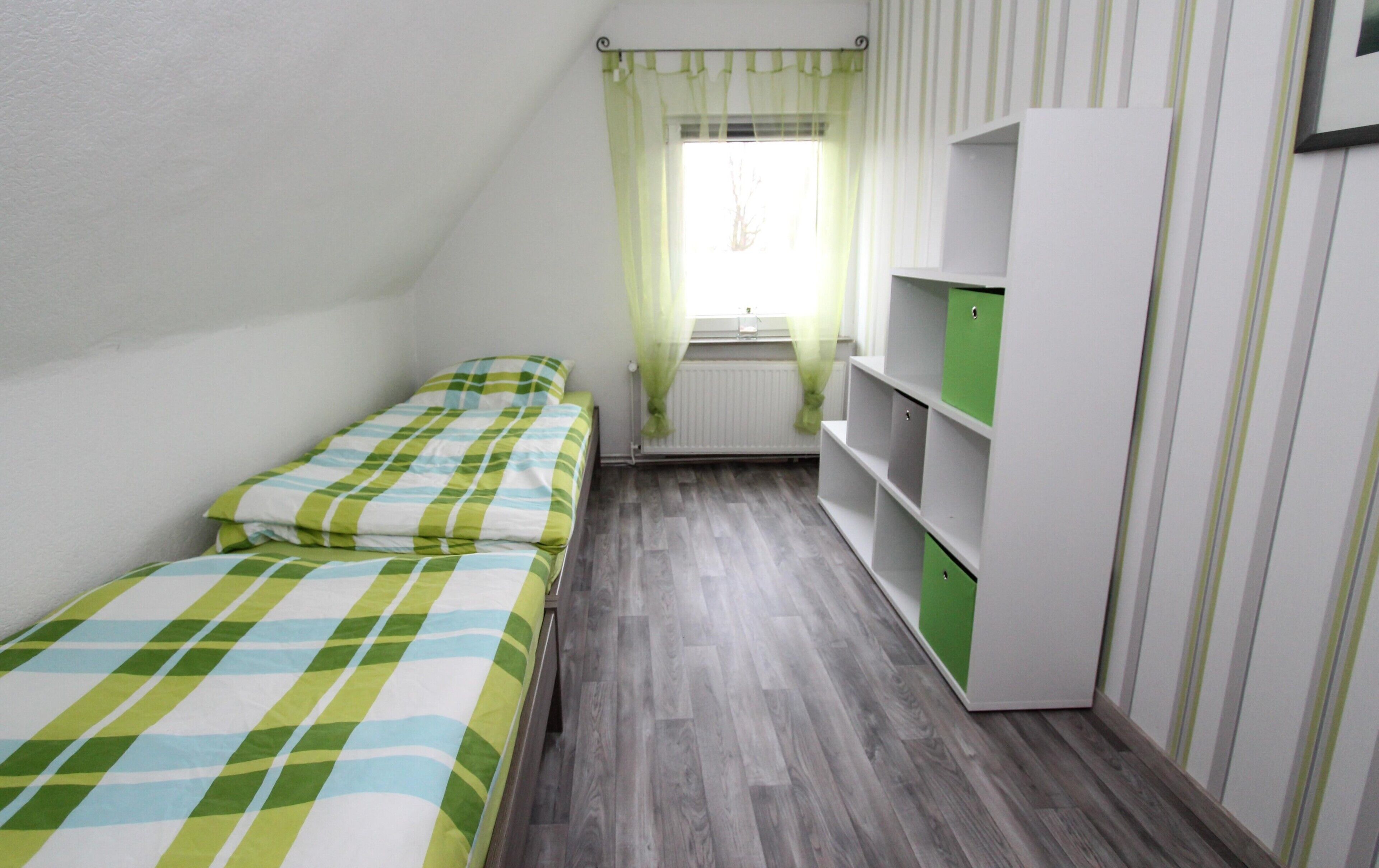 A small room with two single beds, a radiator, a window with sheer curtains, and a white wardrobe with green doors.