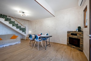 A modern living room with a sofa, dining table, chairs, a fireplace, and a staircase with greenery.