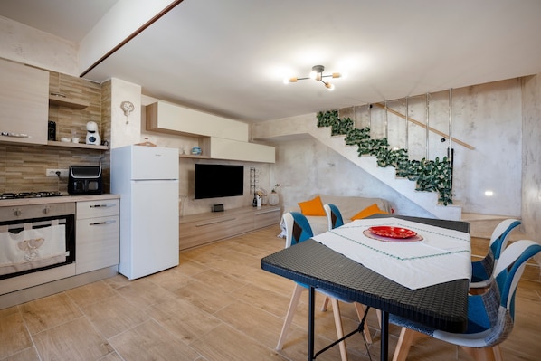 A modern kitchen with a dining area, a flat-screen TV, and a staircase with greenery.