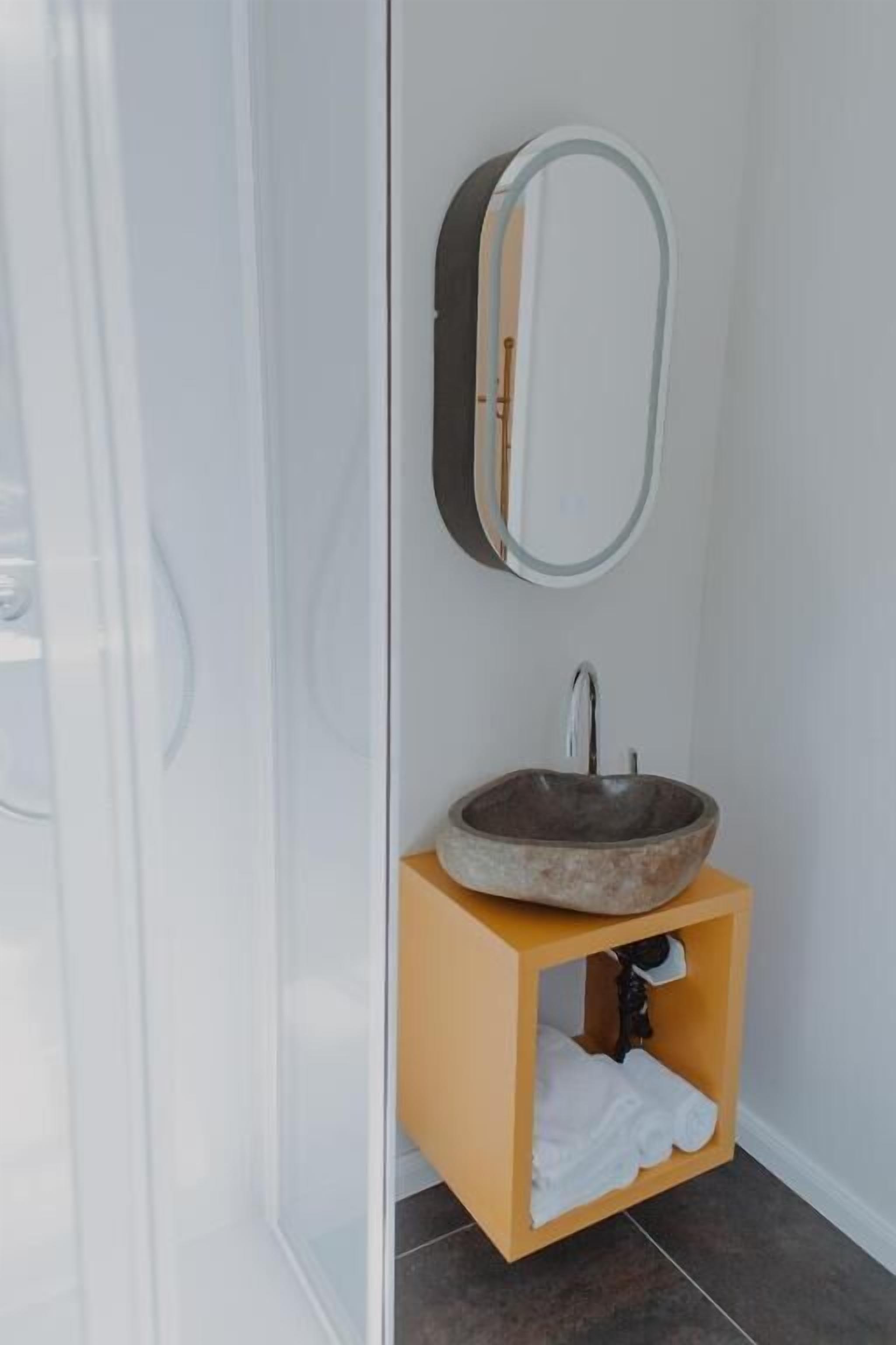 Un baño con cabina de ducha de vidrio, tocador de madera con espejo redondo y lavamanos de piedra.