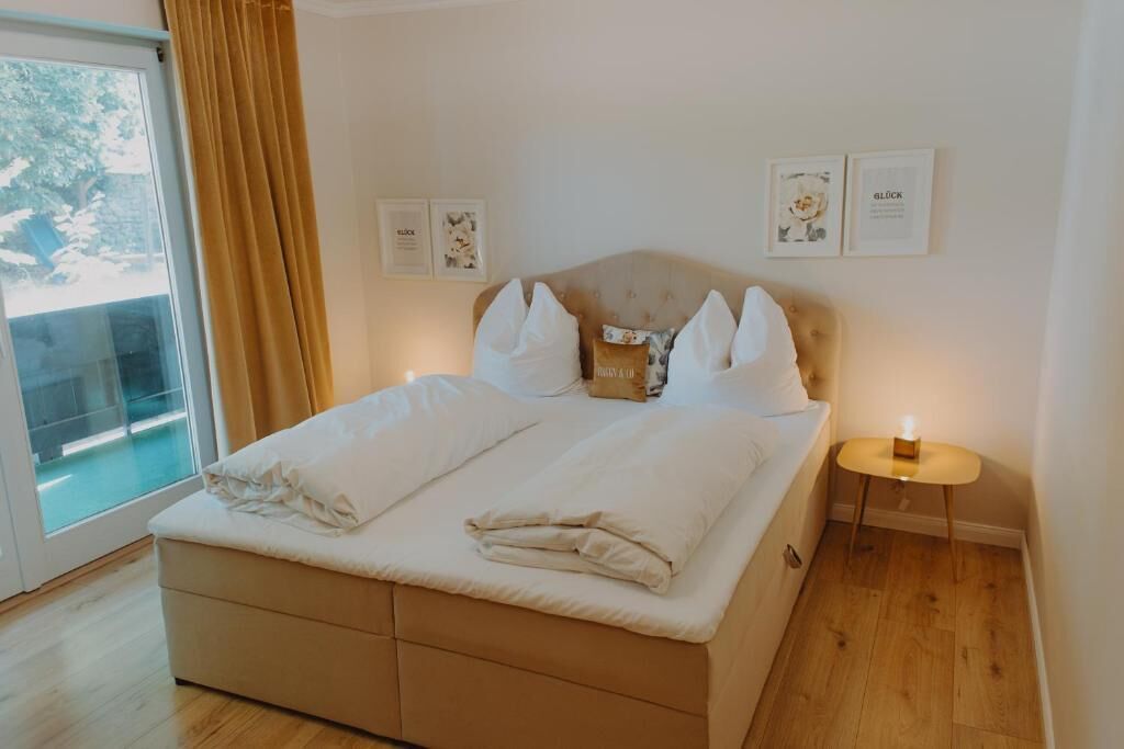 A neatly made bed with white linens and pillows, a bedside table with a lamp, and a view of a balcony through a glass door.