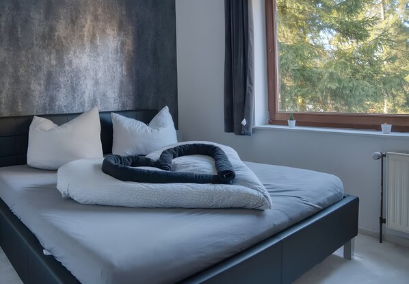 A modern bedroom with a bed, pillows, and a view of trees through a window.