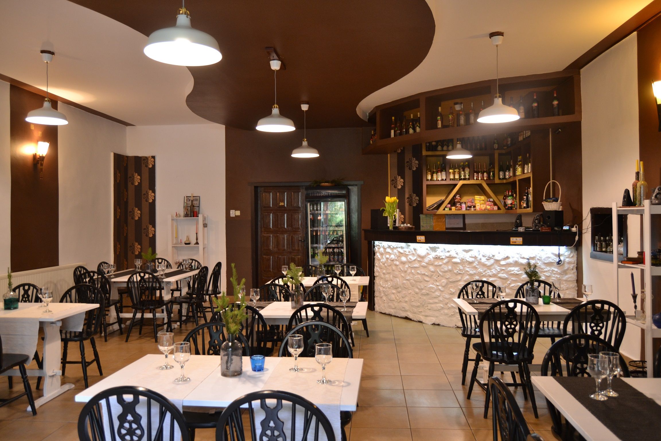 A restaurant interior with multiple dining tables and chairs, pendant lighting, a bar area with shelves of bottles, and a decorative wall.