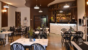 A restaurant interior with multiple dining tables and chairs, pendant lighting, a bar area with shelves of bottles, and a decorative wall.