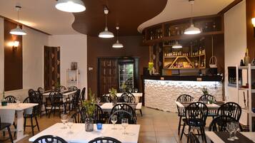 A restaurant interior with multiple dining tables and chairs, pendant lighting, a bar area with shelves of bottles, and a decorative wall.