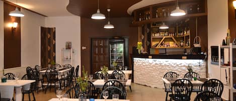 A restaurant interior with multiple dining tables and chairs, pendant lighting, a bar area with shelves of bottles, and a decorative wall.