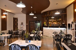 Interior de un restaurante con varias mesas y sillas, iluminación colgante, una zona de bar con estantes llenos de botellas y una pared decorativa.