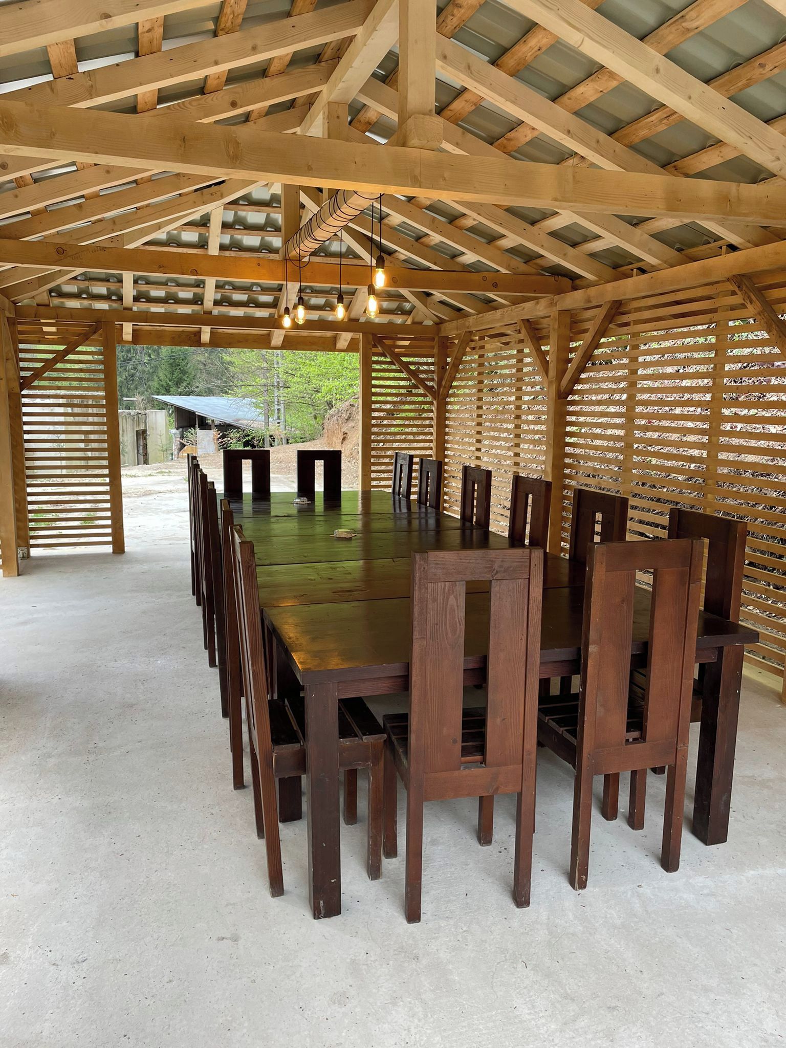 A covered outdoor dining area with a long wooden table and chairs, a concrete floor, and a wooden ceiling structure.