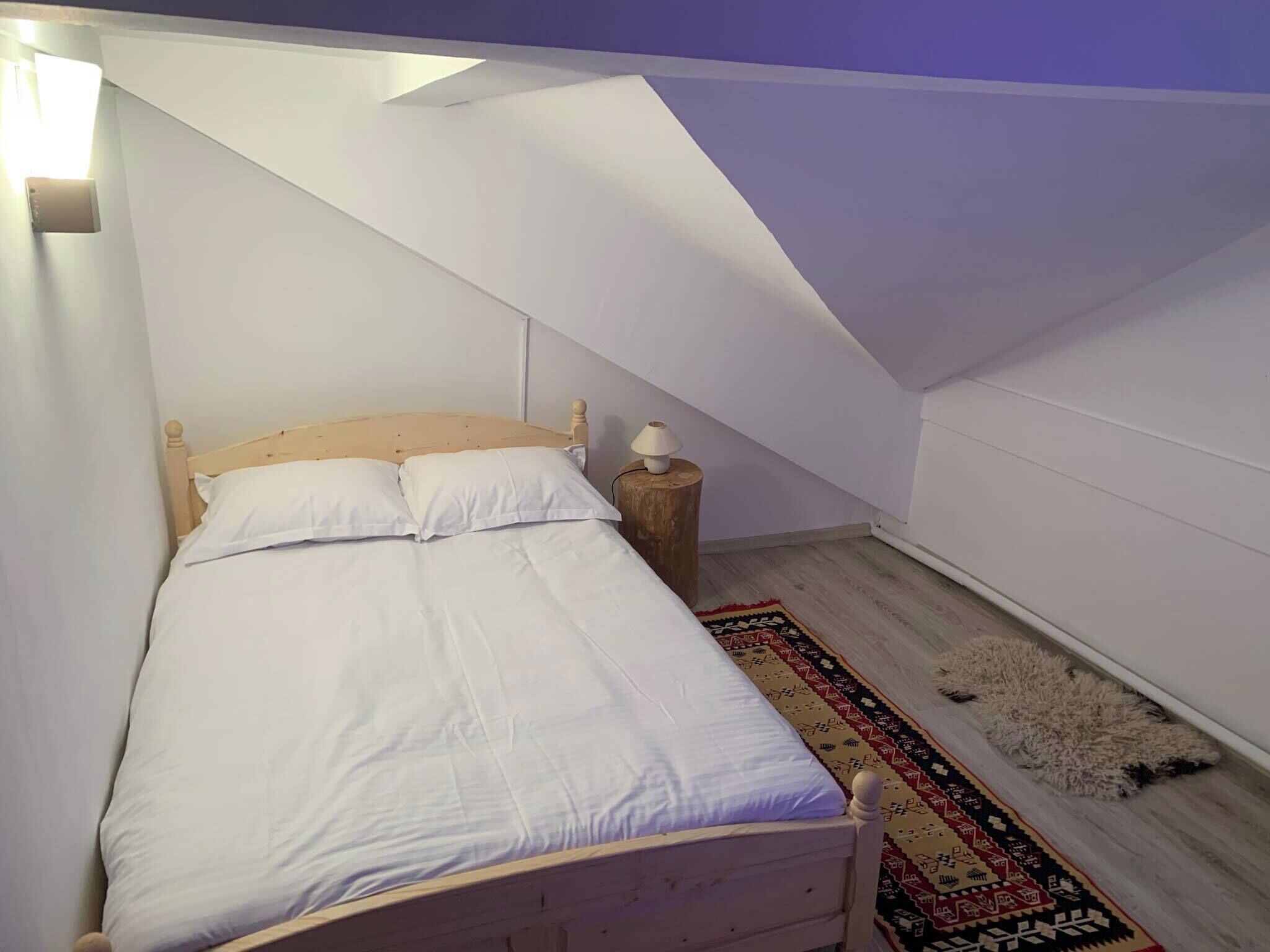 Une chambre avec un lit en bois, une literie blanche, une petite table de chevet avec une lampe et un tapis à motifs sur un plancher en bois.