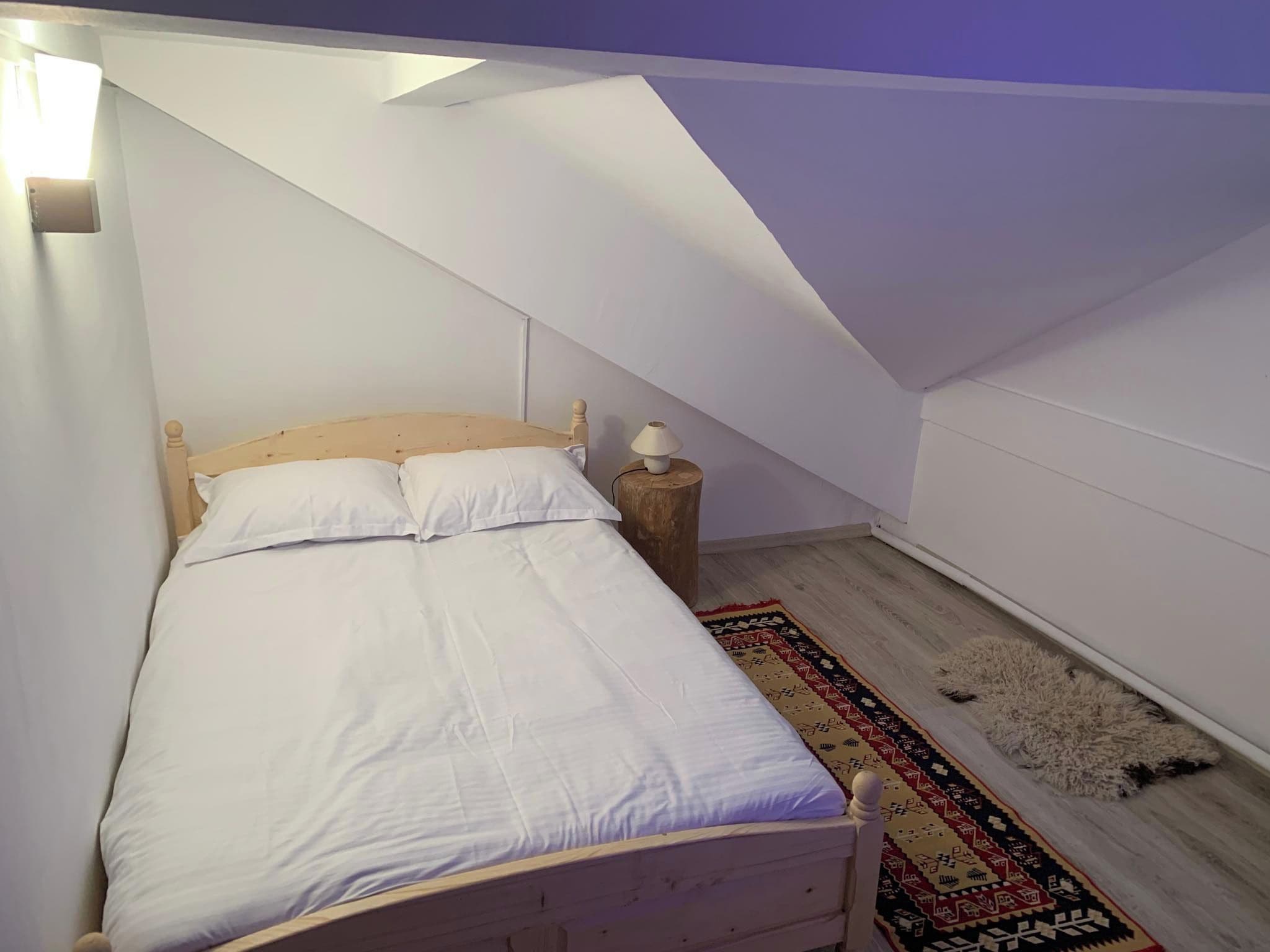 A bedroom with a wooden bed, white bedding, a small bedside table with a lamp, and a patterned rug on a wooden floor.