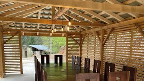 Un área de comedor al aire libre techada, con una larga mesa de madera y sillas, piso de concreto y estructura de techo de madera.