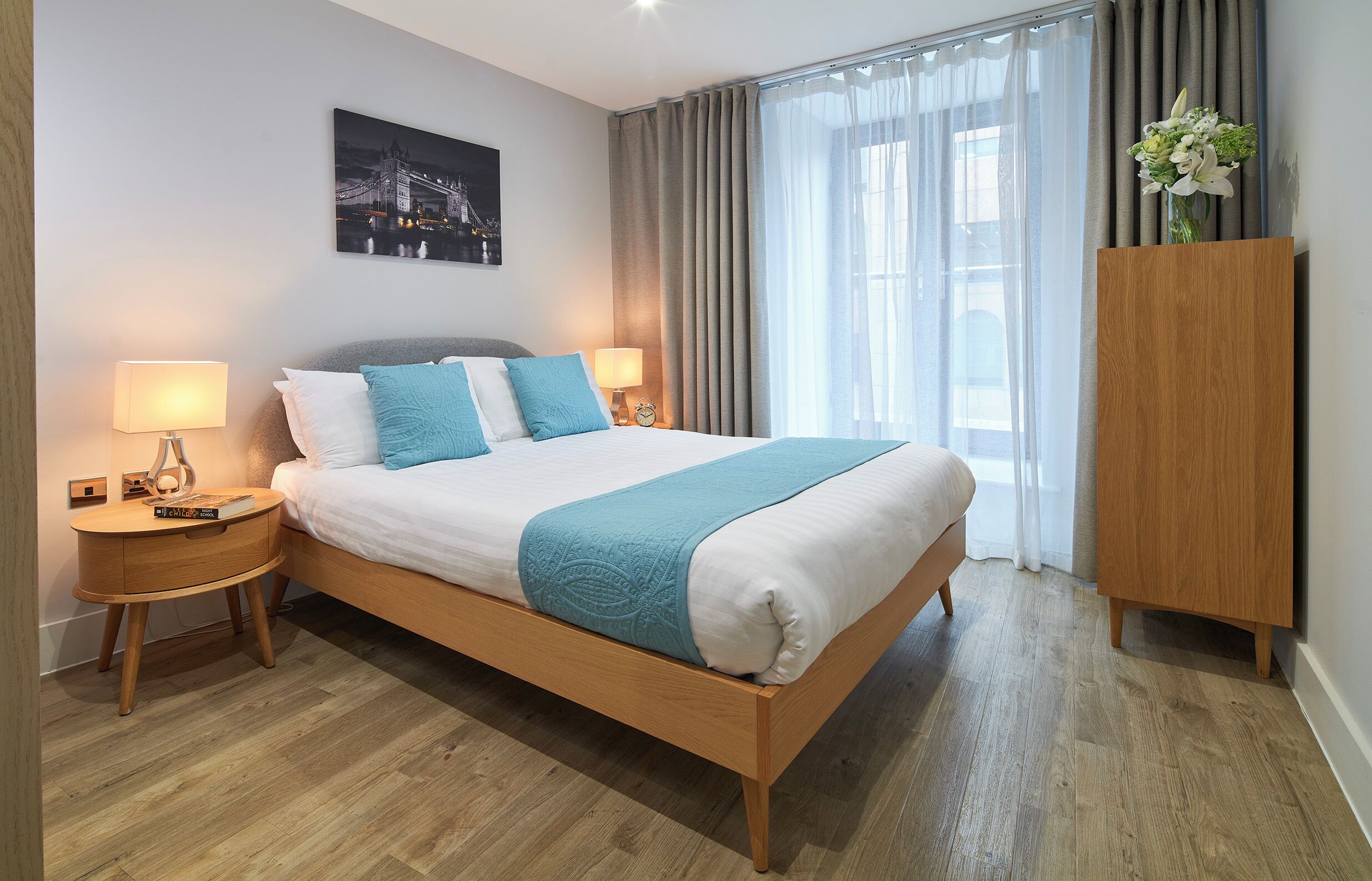 A hotel room with a large bed, wooden nightstands, a night lamp, a framed picture on the wall, and a wooden wardrobe.