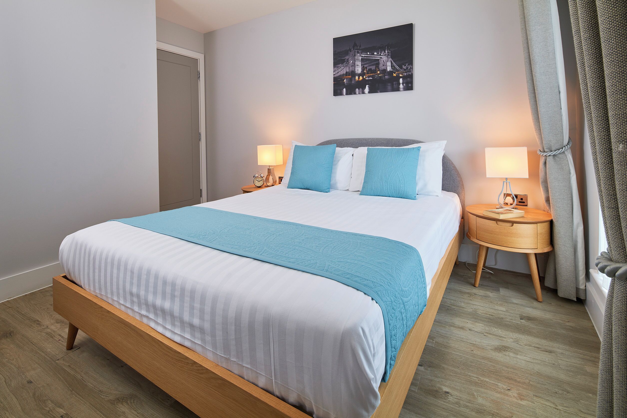 Una cama bien hecha con colcha azul y blanca, dos mesitas de noche con lámparas, suelo de madera y un cuadro colgado en la pared de un puente.