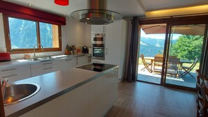 A modern kitchen with a large island, stainless steel appliances, and a view of a mountainous landscape through the sliding glass doors. - Morzine: panoramic, south-facing, comfortable chalet (Morzine)