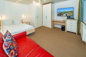 A hotel room with a bed, a red sofa, a desk with a computer and speakers, a TV on the wall, and a door leading to another room. - Haus Conwind apartment 5a - Cozy vacation apartment with sauna, garden and (Sylt-Ost)