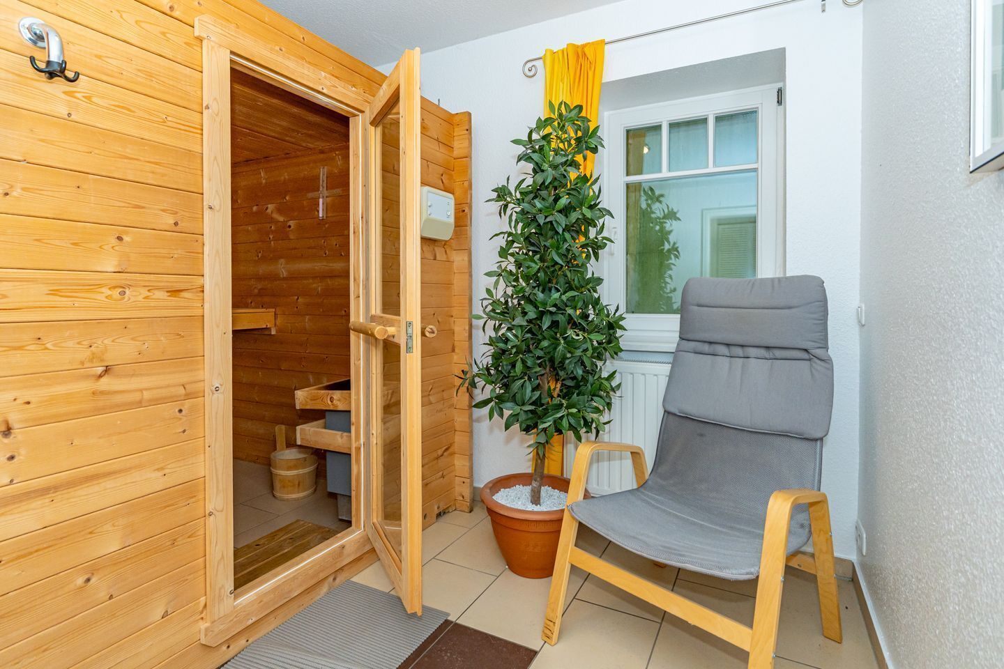 Ein Saunaraum mit Holzwänden, einem grauen Sessel, einem Blumentopf und einem Fenster mit gelben Vorhängen.