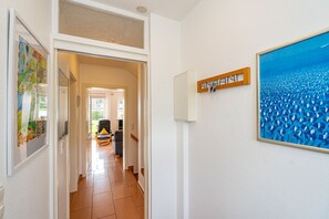 Un couloir avec un sol carrelé, une porte donnant sur une pièce où se trouvent une chaise et une table, et un tableau accroché au mur.