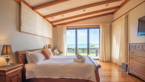 A spacious bedroom with a large bed, wooden headboard, and a view of the sea through large windows. - Tata Tides Beach Front Retreat - Golden Bay at its finest (Tata Beach)