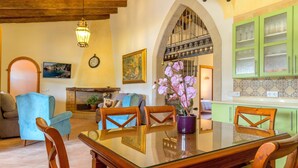 Uma sala de jantar com mesa de vidro e cadeiras de madeira, uma poltrona azul, lareira e armários verdes na cozinha.