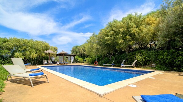 A rectangular swimming pool with lounge chairs and umbrellas, surrounded by trees and a clear sky.