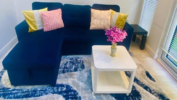 Un sofá azul oscuro con cojines amarillos y rosados, una mesita de noche blanca con un jarrón de flores rosas, y una alfombra de diseño azul y blanco.