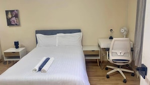 Habitación de hotel con cama, escritorio, silla, mesita de noche, lámpara y ventilador de pared.