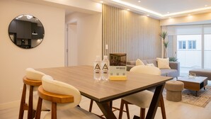 Un espace repas moderne avec une table en bois, des tabourets de bar et un miroir rond. On peut voir deux bouteilles d’eau et une brochure sur la table.