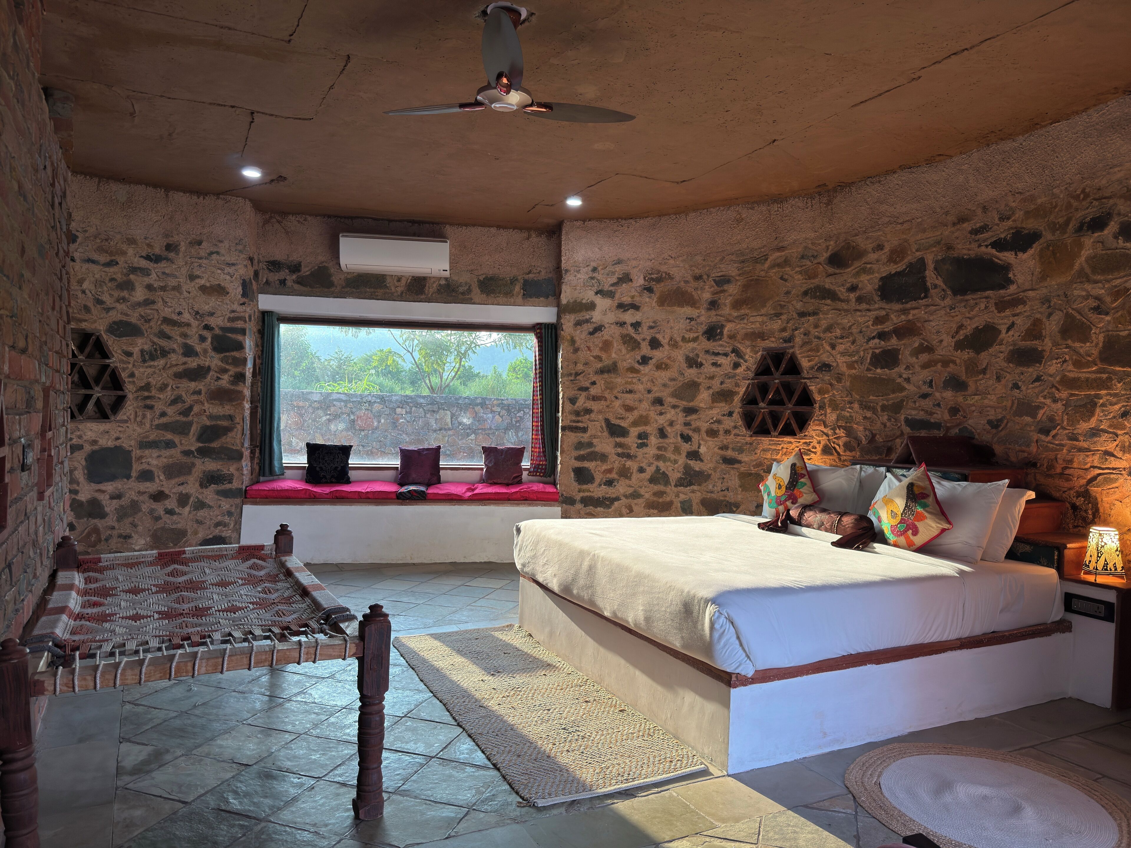 A stone-walled bedroom with a bed, a window with a view, a wooden chair, and a nightstand with a lamp.