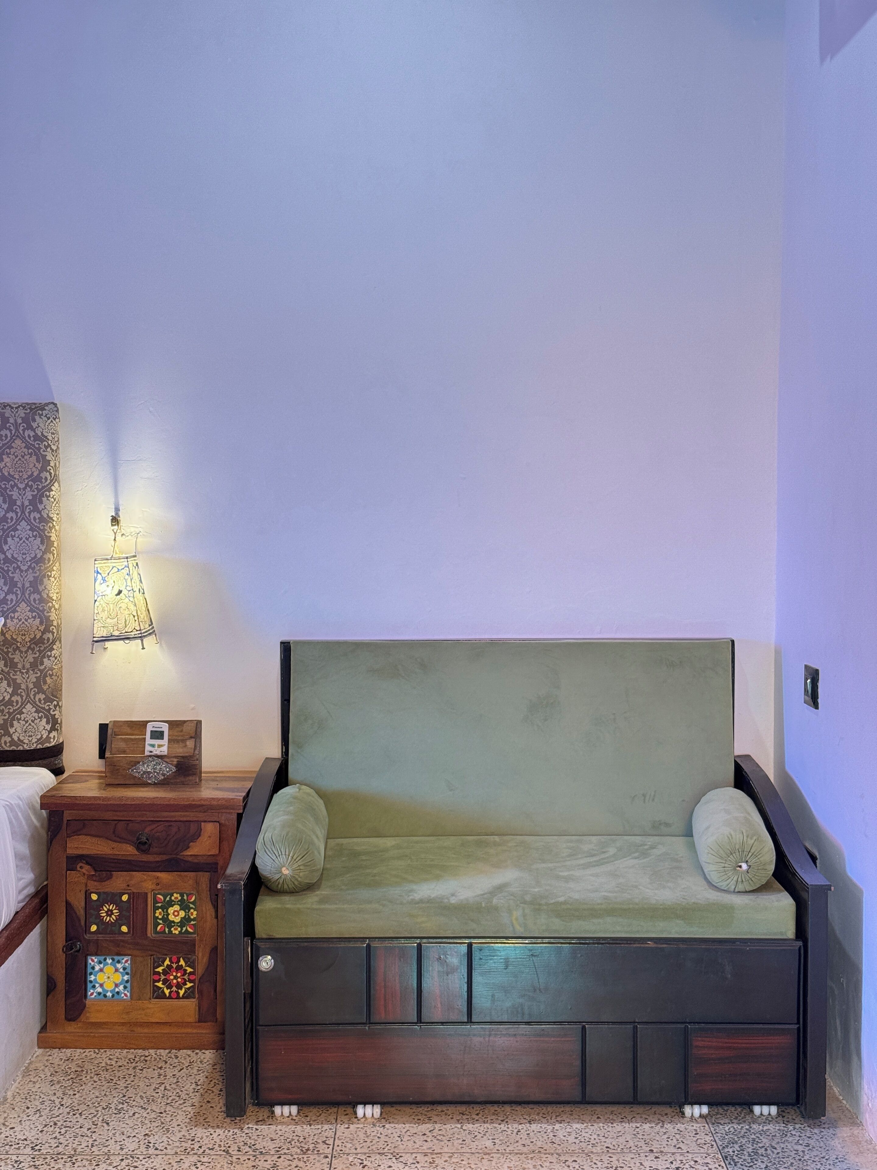 Une petite pièce agréable et chaleureuse, avec un canapé vert, une table de chevet en bois surmontée d’une lampe, et un mur orné d’un motif.