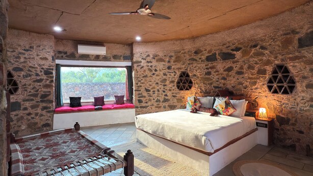 A rustic stone room with a bed, a bench, and a window with a view. - Sariska Kasba - A Village Resort (Rajgarh)
