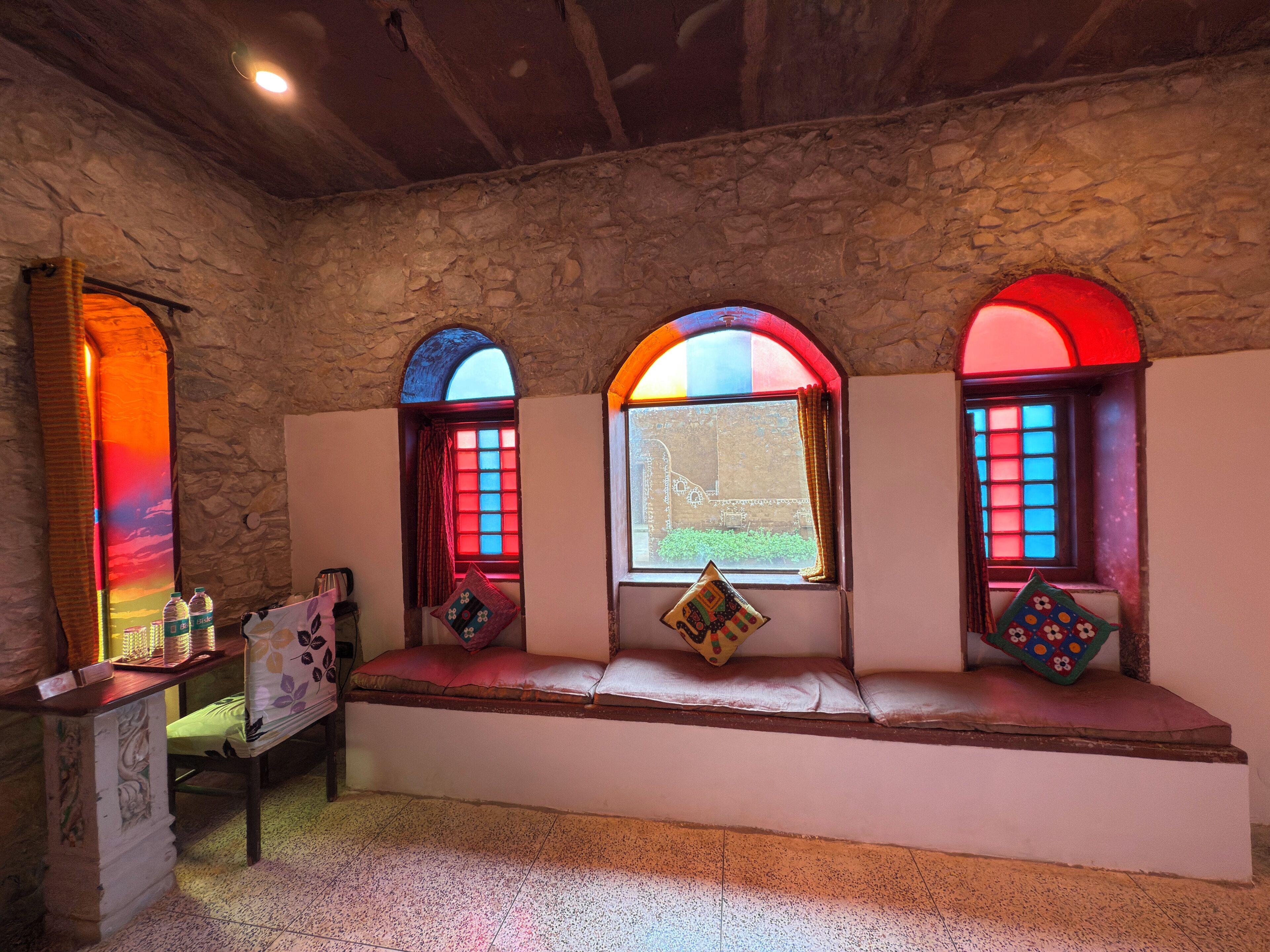A room with stone walls, a bench with cushions, and three arched windows with colorful stained glass.