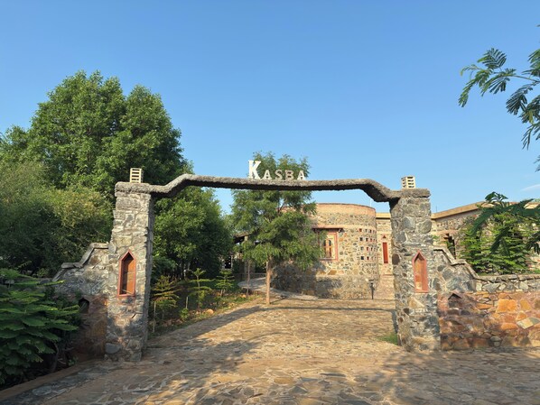A stone archway with the inscription "KASBA" leading to a stone building with arched windows and a circular structure in the background. - Sariska Kasba - A Village Resort (Rajgarh)