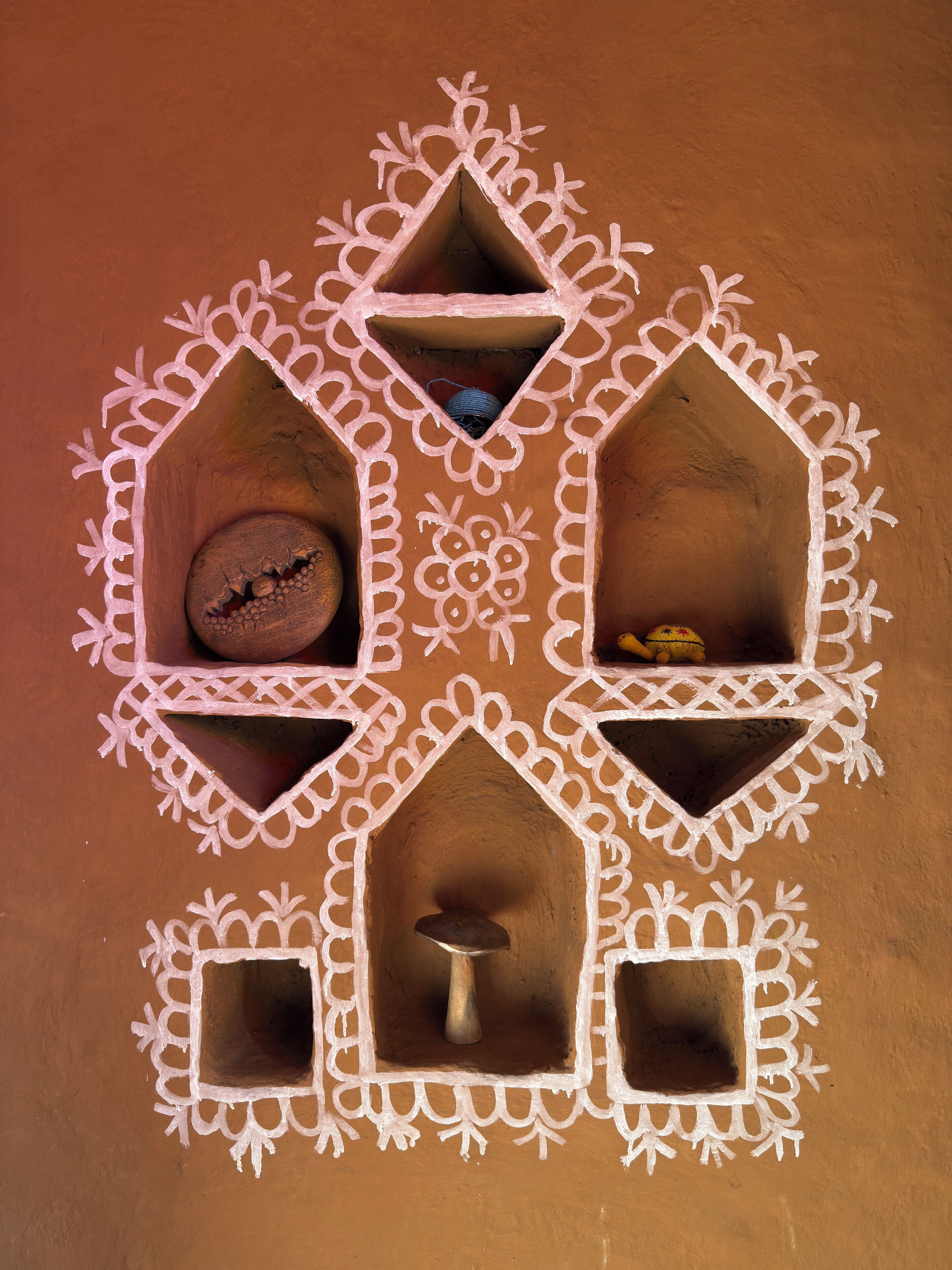 Une maison traditionnelle en argile, ornée de motifs géométriques et comprenant trois petits compartiments, chacun contenant des objets différents.