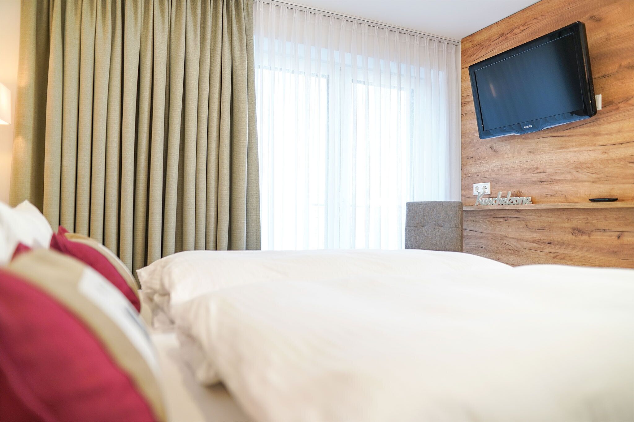 Ein Hotelzimmer mit Bett, einem an einer Holzwand montierten Fernseher und einem Fenster mit Vorhängen.