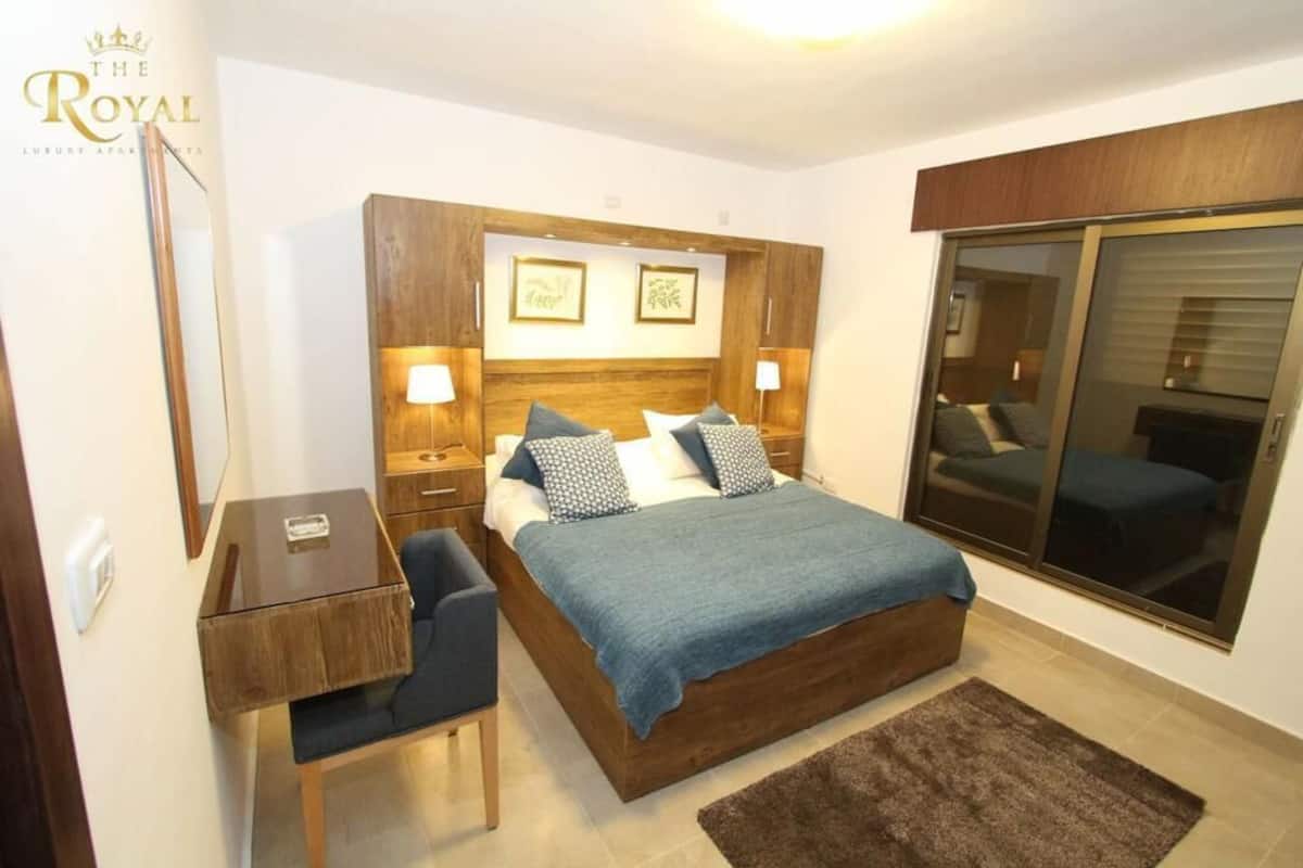 Ein Schlafzimmer mit hölzernem Kopfteil, einem Bett mit blauer Decke, einem Nachttisch, einem Schreibtisch und einem großen Fenster mit Aussicht.