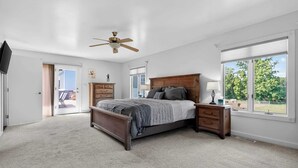 Une chambre à coucher avec un lit, une tête de lit en bois, des tables de chevet, un ventilateur de plafond et une grande fenêtre donnant sur des arbres.