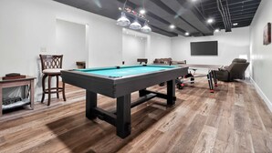 Une table de billard dans une pièce au plancher en bois, avec des tuyaux apparents au plafond et un téléviseur fixé au mur.