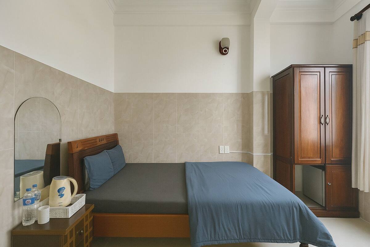 A bedroom with a bed, a wooden wardrobe, a bedside table with a lamp, and a mirror.