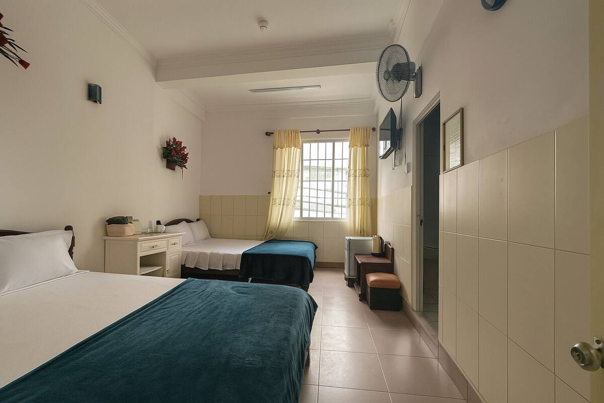 A hotel room with two beds, a desk, a chair, a small table, a fan, and a window with curtains.