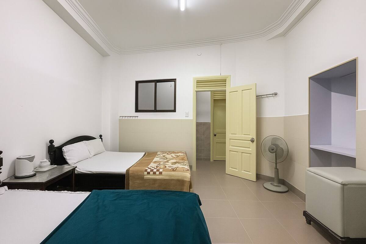 A hotel room with a bed, bedside table, a small table with a kettle, a fan, and a wardrobe.