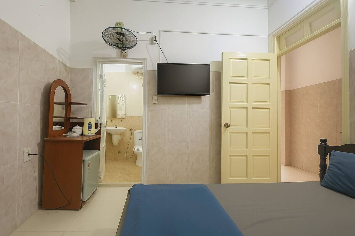 A bedroom with a bed, a dresser, a TV, a fan, and a door leading to a bathroom.