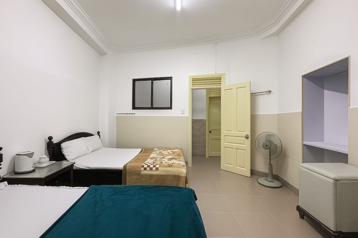 A hotel room with a bed, bedside table, a small table with a kettle, a fan, and a wardrobe.