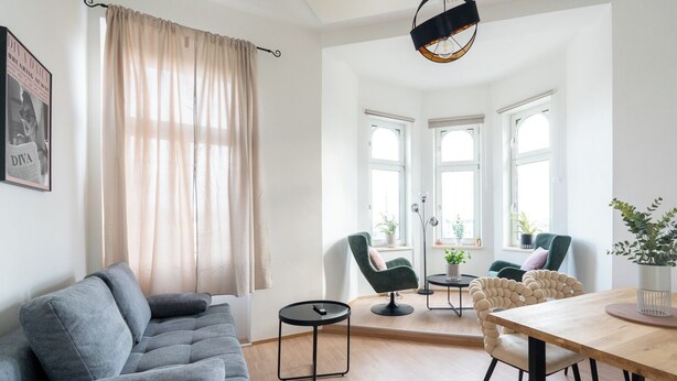 Un salon moderne avec un canapé gris, une table à manger en bois et deux chaises. Il y a trois fenêtres qui laissent entrer la lumière naturelle, un lampadaire et un tableau encadré accroché au mur.