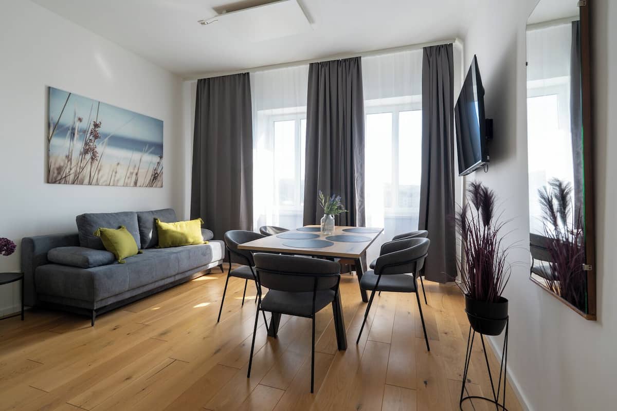 Ein modernes Wohnzimmer mit grauem Sofa, einem Esstisch mit Stühlen, einem wandmontierten Flachbildfernseher und großen Fenstern mit grauen Vorhängen.